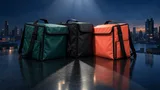 Three unbranded insulated delivery courier bags in green, black, and coral arranged on a reflective dark table, two overlapping closely to symbolize a possible merger, lit dramatically against a moody dark background.