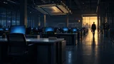 A nearly empty corporate office at dusk with rows of unoccupied desks under blue monitor glow, a chip-shaped sculpture hanging overhead, and a lone figure walking toward a lit exit.
