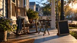 A single wooden porch rocking chair sits beside a small folding table holding a ceramic mug of coffee and a stack of coins, with a "Room for Rent" placard leaning against a nearby lamppost outside a modest house.