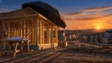 An unfinished wooden house frame at dusk in a suburban development, with a large open black umbrella leaning against it and rows of finished homes stretching into the background under a warm sunset sky.