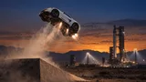 A white sports car flipping in mid-air with visible underbody thrusters above a desert rocket test site at dusk, with a distant launch tower silhouette.